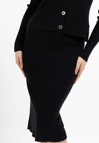 Costum două piese din tricot negru cu creeor, care include un top ajustat cu mânecă lungă și știfturi aurii și o fustă creion cu un tiv evazat.