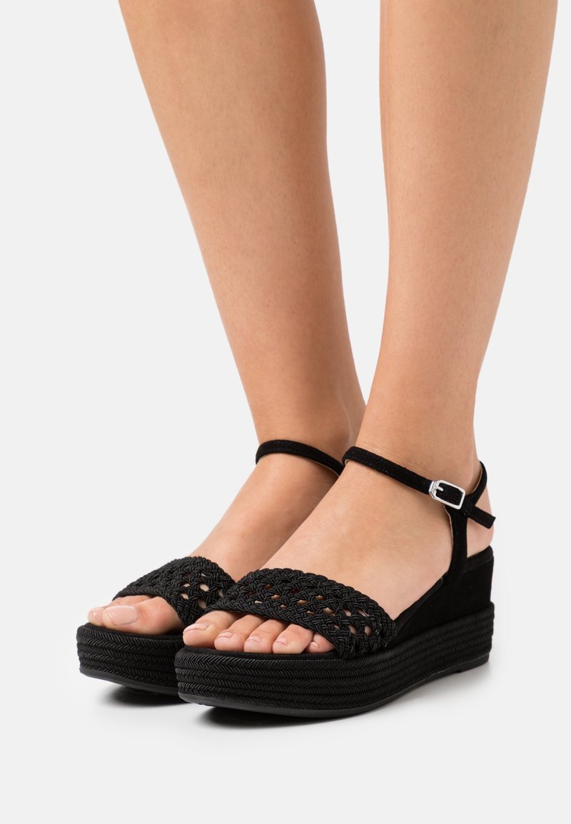 Pieds portant des sandales compensées noires avec des brides tissées à l'avant et des boucles à la cheville sur fond blanc.