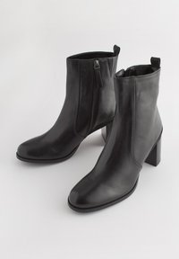 Černé kožené kotníkové boty s elegantním designem, špičatou špičkou a blokovým podpatkem. Obsahují boční zip a poutko pro snadné obouvání.