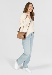 Borsa a tracolla in pelle color cuoio con patta magnetica, decorata con logo a forma di ancoraggio, cinturino regolabile, abbinata a un maglione crema e jeans azzurri chiari.