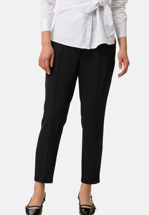 Schwarze, taillierte Hose mit slim fit, hoher Taille und vorderen Falten. Kombiniert mit glänzenden schwarzen Ballerinas und einem weißen Hemd.