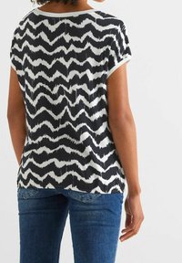 Černobílé tričko s krátkým rukávem a vzorem ve tvaru chevronu, s kulatým výstřihem, vyrobené z hladké tkaniny, v kombinaci s modrými džíny.
