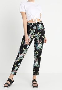 Pantalon noir à motif floral avec des feuilles vertes et des fleurs roses, associé à un haut court blanc avec un nœud à l'avant. Sandales plates noires.