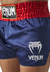 Pantalones cortos atléticos Venum en satén azul marino, con una cinturilla elástica roja y un logo blanco. Textura suave con acentos de color en contraste.