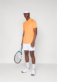 Oransje polo skjorte med krage, hvite shorts, hvite joggesko og en hvit caps. Holder et tennistrekk og en tennisball.