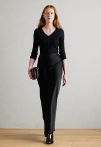 Top en tricot noir à côtes avec un col en V et des manches trois-quarts, associé à une jupe noire ajustée. Le mannequin tient une petite pochette noire et porte des bottines noires.