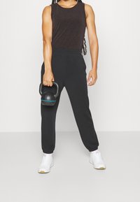 Pantaloni jogging neri con polsini elastici e vita alta, abbinati a una canotta aderente marrone scuro. Scarpe sportive bianche e una kettlebell in mano.