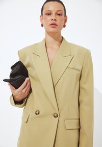 Béžová dvojradová sako s ostrými chlopňami, dvoma prednými vreckami a gombíkmi v imitácii želvoviny. Modelka drží čiernu textúrovanú kabelku.