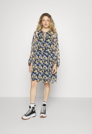 Vestido floral com fundo azul marinho, mangas longas e decote com laço; combinado com ténis de cano alto pretos e meias brancas com riscas.
