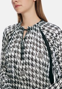 Schwarz-weiße gemusterte Bluse mit Rüschenausschnitt, halbem Reißverschluss und schwarzen Akzenten an den Seiten, mit einem Logo.