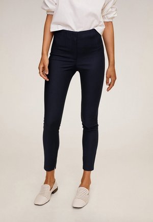 Femme portant un pantalon noir slim, un haut blanc à manches bouffantes et des chaussures blanches tissées slip-on, debout sur un fond clair uni.