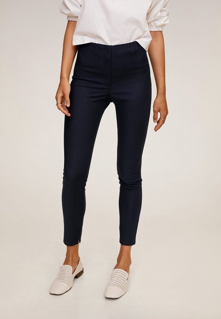 Femme portant un pantalon noir slim, un haut blanc à manches bouffantes et des chaussures blanches tissées slip-on, debout sur un fond clair uni.