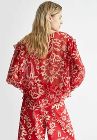 Blusa rossa con motivo floreale in tessuto trasparente, maniche a volant e pantaloni abbinati. Presenta intricate decorazioni floreali bianche.