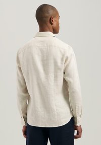 Lininė marškiniai off-white spalvos, ilgomis rankovėmis, sagstomais manžetais ir šiek tiek suapvalintu apatine dalimi. Lygus tekstūra ir kasdieniškas apykaklės dizainas.