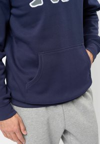 Sudadera azul marino con bolsillo frontal y bordado tonal, combinada con pantalones de chándal gris claro. La tela parece suave y con textura.