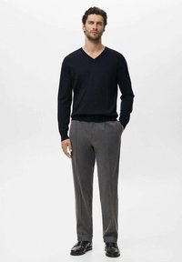 Suéter de color azul marino oscuro con escote en V, combinado con pantalones grises con pliegues y zapatos negros, de pie frente a un fondo blanco liso.