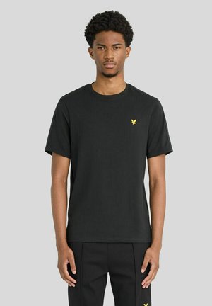 Junger Mann, der nach vorne schaut, trägt ein einfaches schwarzes T-Shirt mit einem kleinen gelben Vogel-Logo auf der Brust und schwarze Hosen.