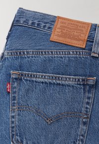 Jeans en denim bleu moyen avec une étiquette en cuir sur la ceinture marquée "LEVI STRAUSS & CO." Étiquette rouge sur la poche arrière, détails de couture classiques.