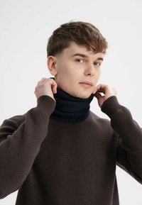 Bruine gebreide trui met een donkerblauwe ribgebreide col. Model past de col aan, waardoor de gestructureerde stof en de aansluitende pasvorm zichtbaar worden.