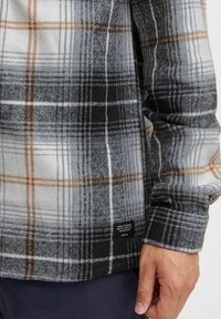Plaid flanelinė marška, kurioje dominuoja pilka, juoda ir smėlio spalvos, su minkšta tekstūra ir etiketė apačioje. Ilgos rankovės, laisvas siluetas.