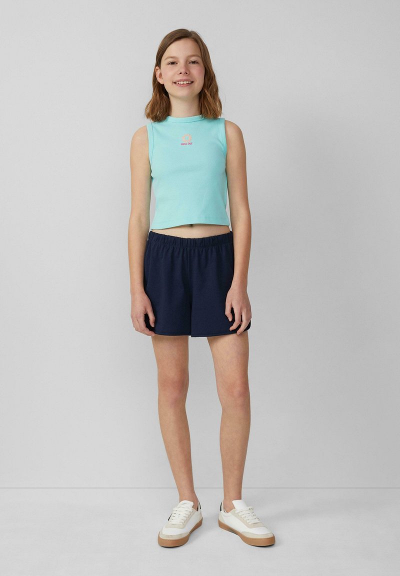 Lichtblauw mouwloos crop top met logo, gecombineerd met marineblauwe shorts met elastische taille en witte sneakers met beige accenten. Eenvoudige en casual outfit.