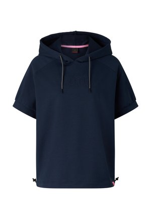 Blondynka ubrana w oversize granatową bluzę z kapturem z krótkim rękawem, pasujące spodnie dresowe, białe sneakersy i duże lustrzane okulary przeciwsłoneczne, stoi zwrócona twarzą do przodu.