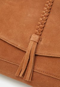 Primo piano di una borsa in suede marrone con una tracolla intrecciata e un dettaglio a nappina sul risvolto anteriore.