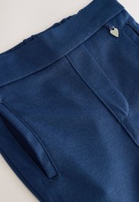 Pantalones azul marino con textura suave, cintura elástica y bolsillos laterales. Presentan un pequeño adorno metálico en forma de corazón en la cinturilla.