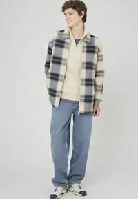 Veste à carreaux beige et marine sur un pull beige clair, associée à un jean bleu ample et des baskets grises. Style décontracté, coupe relax et superpositions.