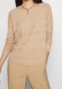 Beige stickad tröja med rund halsringning, långa ärmar och en något krökt fåll. Ytan är slät med subtila horisontella ränder.