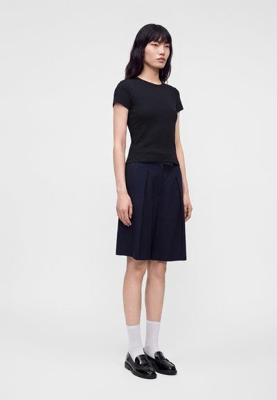 Femme debout portant un haut noir à manches courtes, une jupe bleu marine longueur genoux, des chaussettes blanches et des mocassins noirs sur un fond clair uni.