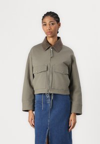 Giacca verde oliva tagliata con colletto in suede marrone, zip frontale e due tasche sul petto, abbinata a una gonna in denim con uno spacco.