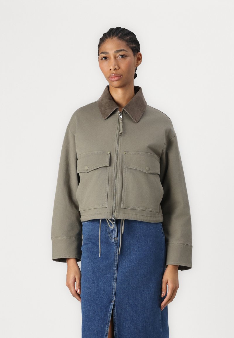 Giacca verde oliva tagliata con colletto in suede marrone, zip frontale e due tasche sul petto, abbinata a una gonna in denim con uno spacco.