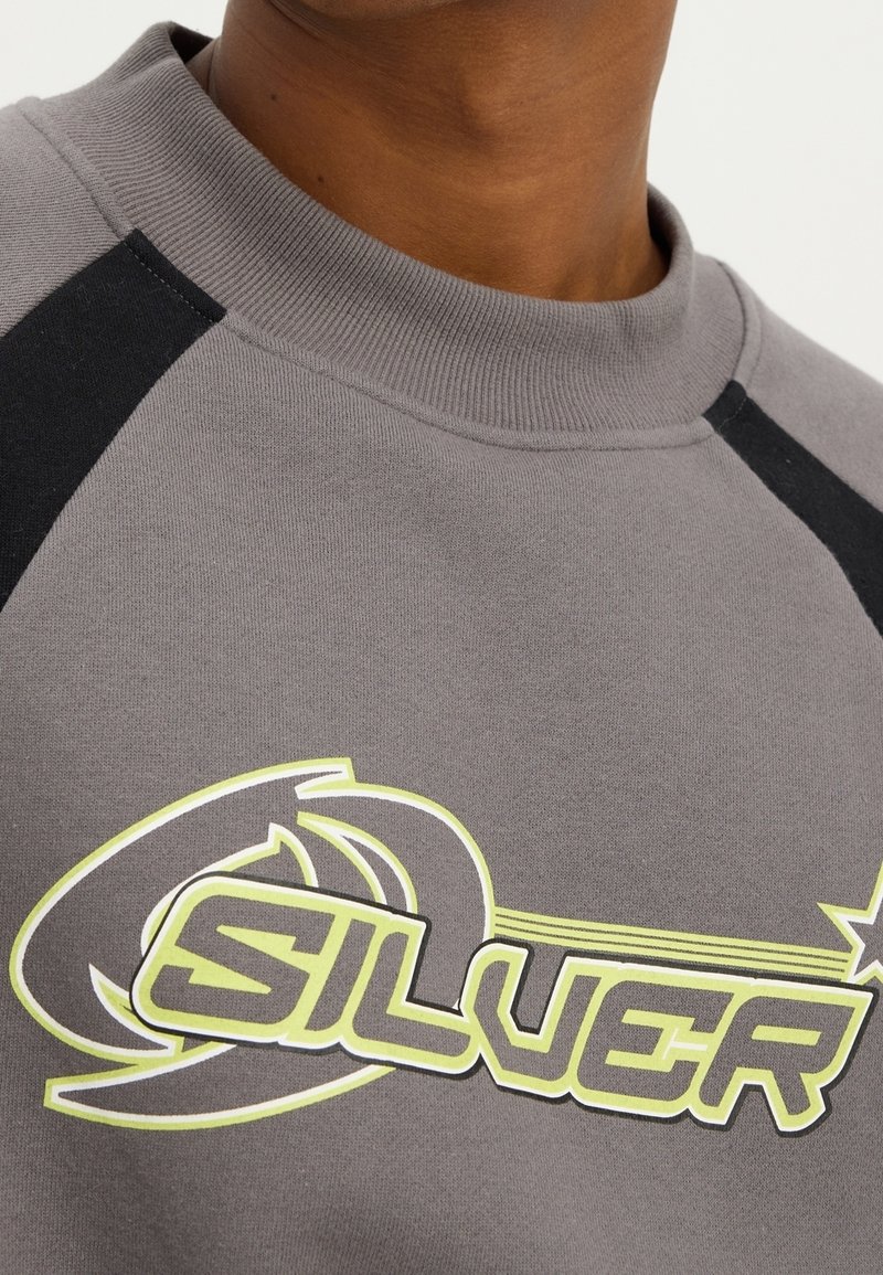 Szara bluza z czarnymi akcentami, z wyraźnym napisem "SILVER" w neonowej żółci i czerni. Miękka faktura, okrągły dekolt i raglanowe rękawy.