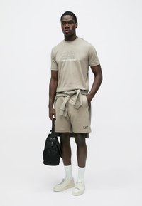 Μπεζ βαμβακερό t-shirt με το λογότυπο EA7, σετ με αντίστοιχα σορτσάκια. Λευκές κάλτσες και αθλητικά παπούτσια. Μαύρη τσάντα κρατημένη σε ένα χέρι.