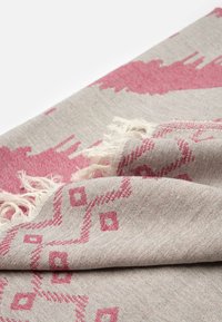 Textil tejido con un fondo gris claro y patrones geométricos en rosa, con bordes con flecos, que muestra un acabado texturizado.