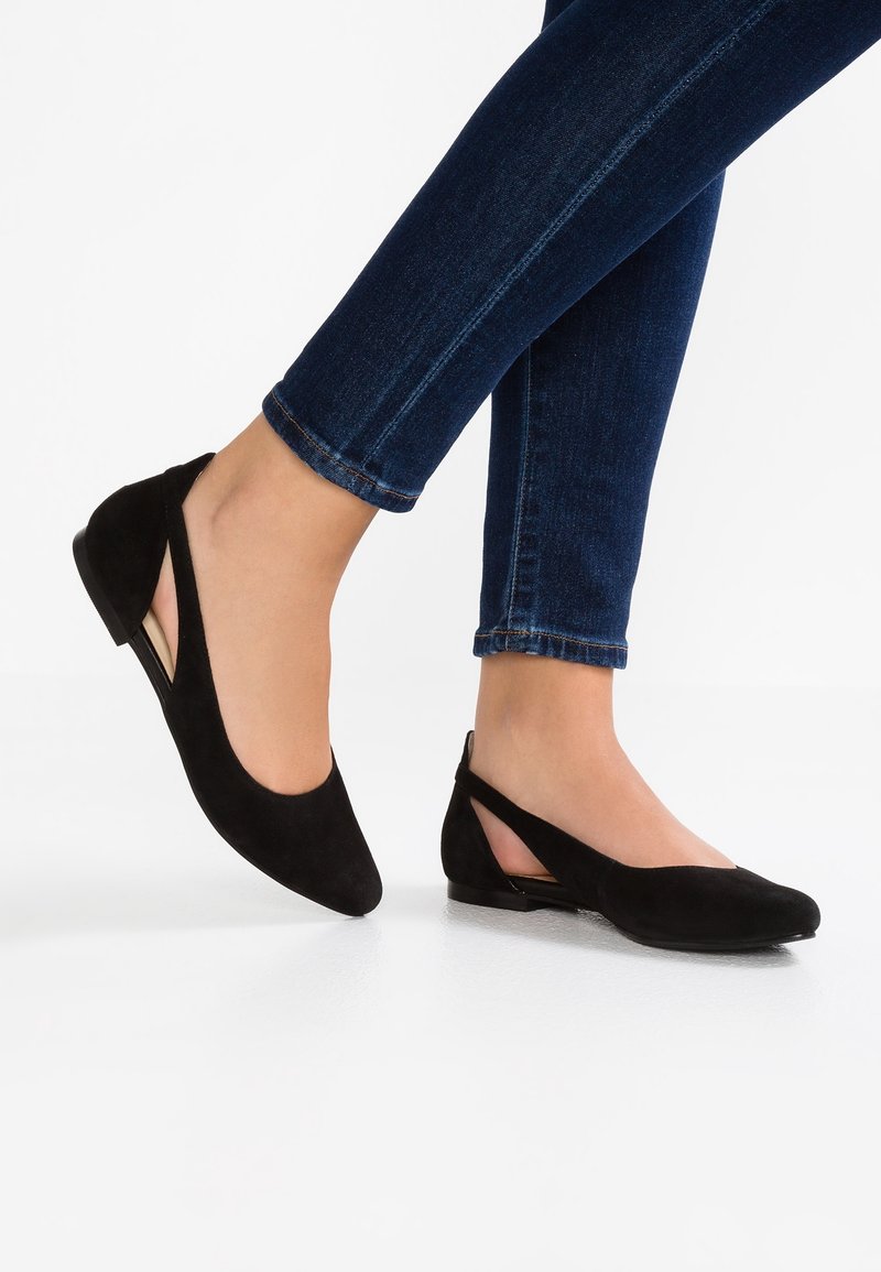 Schwarze Wildleder-Ballerinas mit spitzem Zehenbereich, ausgeschnittenen Seiten und flachem Absatz. Kombiniert mit dunkelblauen Jeans, die ihr elegantes Design zur Geltung bringen.