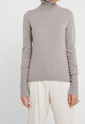 Woman wearing a light gray ribbed turtleneck sweater and loose-fitting white pants, with gold rings on her right hand.