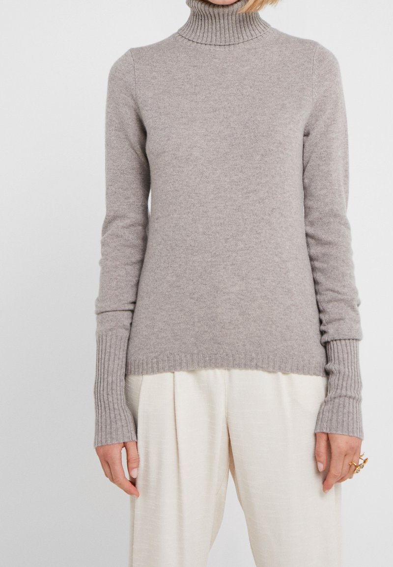 Femme portant un pull à col roulé côtelé gris clair et un pantalon ample blanc, avec des bagues en or à la main droite.