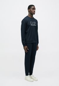 Muž stojící vpravo, oblečený v tmavě modré mikině a odpovídajících teplácích s bílými teniskami na jednoduchém světlém pozadí.