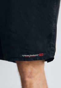 Black denim shorts with a crinkled texture. Features a red and white "Diesel Industry" label on the hem. Stitched edges are visible.