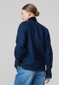 Camicia di jeans blu scuro con maniche lunghe, polsini abbottonati e dettaglio sulla cucitura posteriore. Abbinata a pantaloni di jeans blu chiaro.