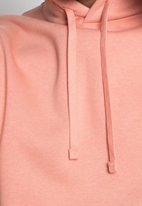 Hoodie i ljus korall, med en mjuk struktur, dragsko och en avslappnad passform. Detaljen betonar tyget och sömmen.