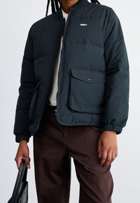 Giacca puffo verde scuro con zip frontale, colletto a costine e polsini elasticizzati; presenta una tasca a toppa e un piccolo logo "OBEY".