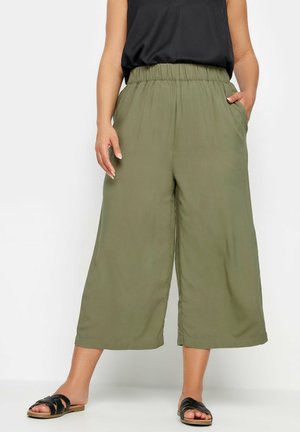 Olivgrüne weite Hosen mit einem elastischen Taillenbund, seitlichen Taschen und einer glatten Stofftextur, kombiniert mit schwarzen Riemchensandalen.