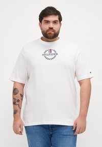 Balta medvilninė T-shirt marškinėliai su trumpomis rankovėmis, priešpusėje su mėlyna ir raudona „Hilfiger“ logotipu bei laurelo dizainu. Laidų stilius.