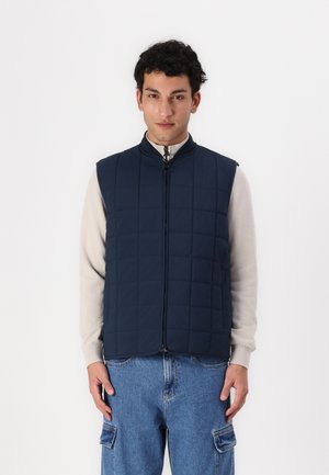 Gepunktete marineblaue Weste mit hohem Kragen und Reißverschluss auf der Vorderseite. Getragen über einem hellen Langarmshirt, kombiniert mit blauen Cargo-Hosen.