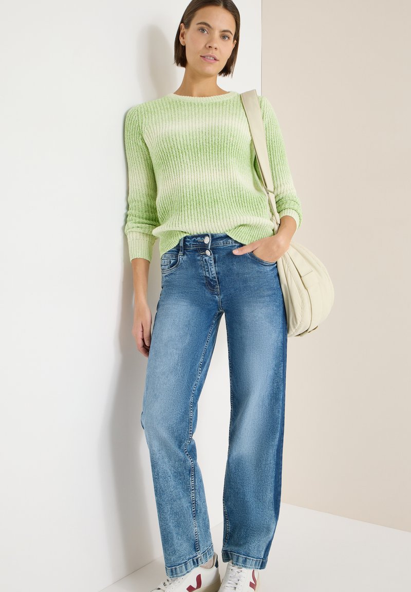 Pull tricoté rayé vert clair, jeans larges bleus, et baskets blanches avec des accents rouges. Un sac beige pend de l'épaule.