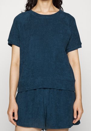 Vrouw draagt donkerblauw kort mouw t-shirt en bijpassende losse shorts, staand met ontspannen armen langs het lichaam tegen een effen achtergrond.