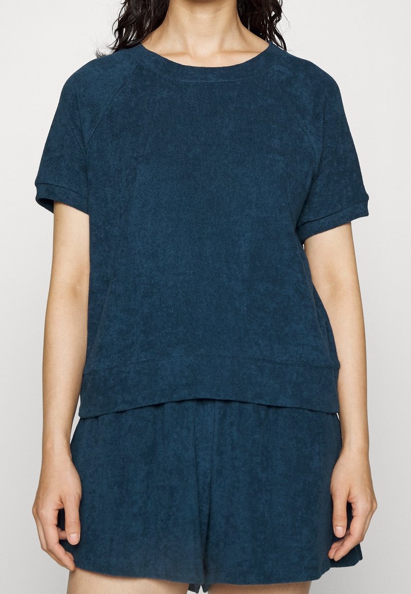 Femme portant un haut à manches courtes bleu foncé et un short ample assorti, debout les bras détendus le long du corps, sur un fond uni.